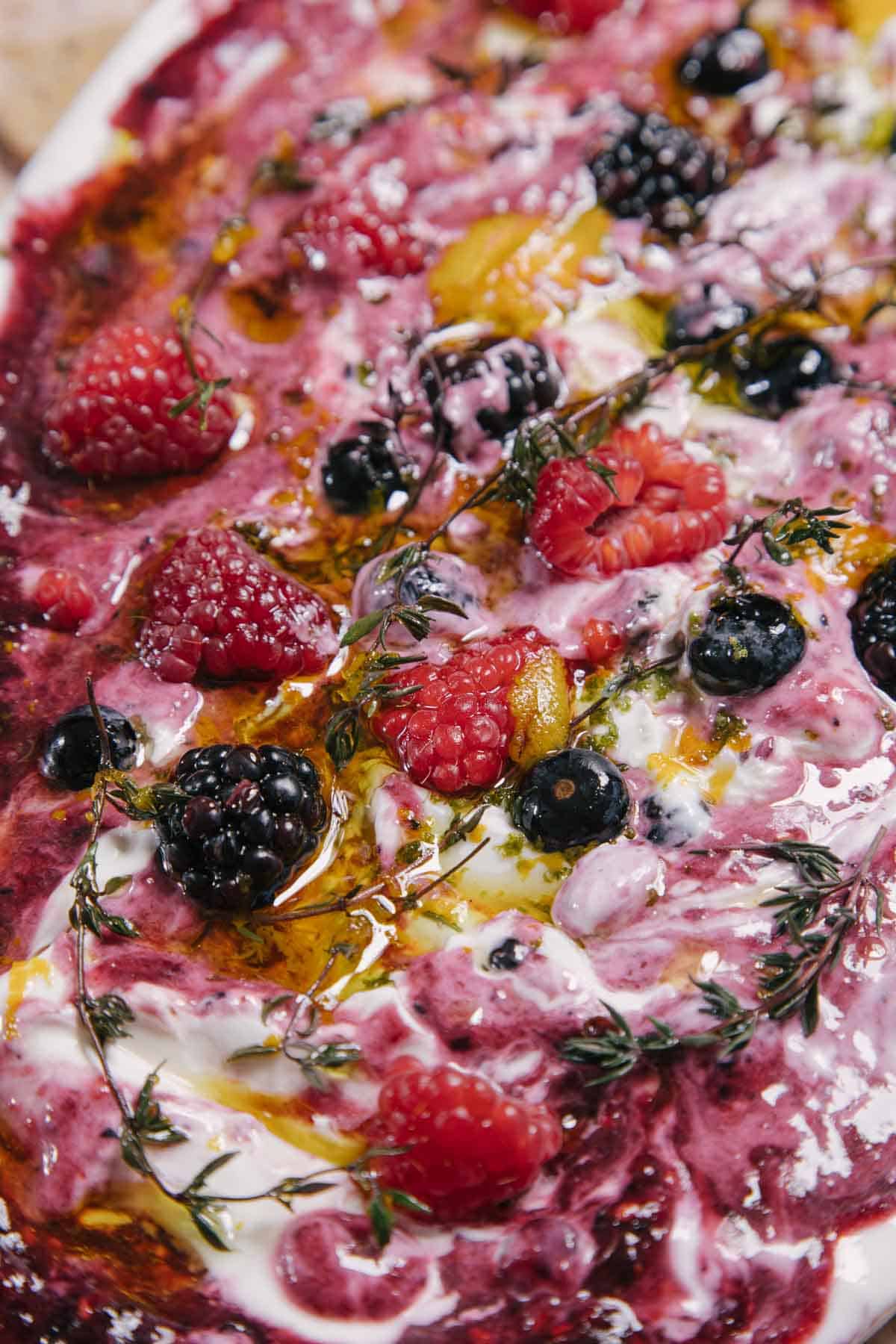 A close-up of a vibrant dish featuring swirls of yogurt with various berries, including raspberries, blackberries, and blueberries. Sprigs of thyme are scattered on top, and a drizzle of olive oil adds a glossy finish.