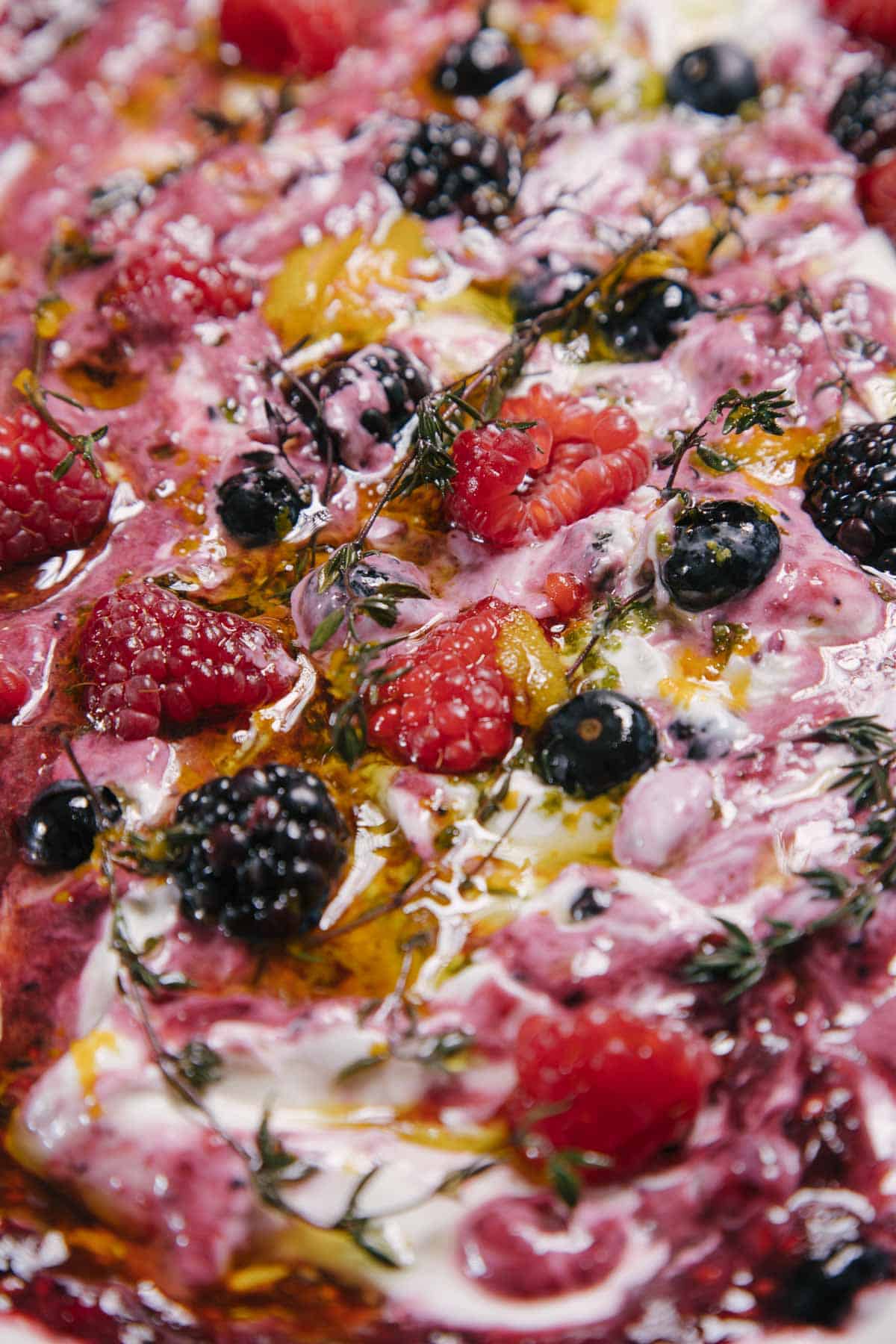 Close-up of a colorful dish featuring a variety of berries like raspberries, blackberries, and blueberries mixed with creamy yogurt, garnished with fresh herbs. The dish has a textured appearance with a mix of vibrant reds, purples, and whites.