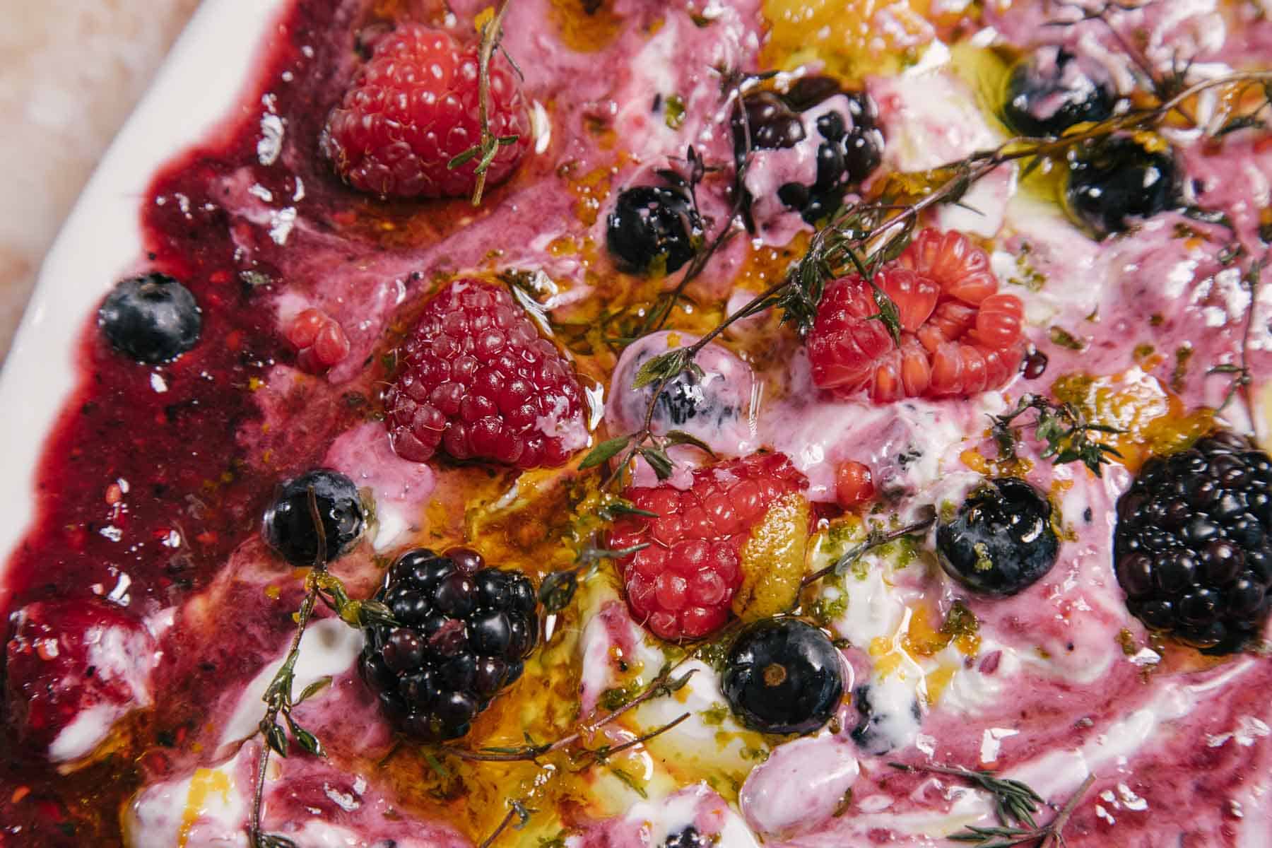 Close-up of a colorful dessert featuring a swirl of red, pink, and white creams topped with fresh raspberries, blackberries, blueberries, and sprigs of thyme. The mixture has a glossy appearance with a slightly uneven texture.