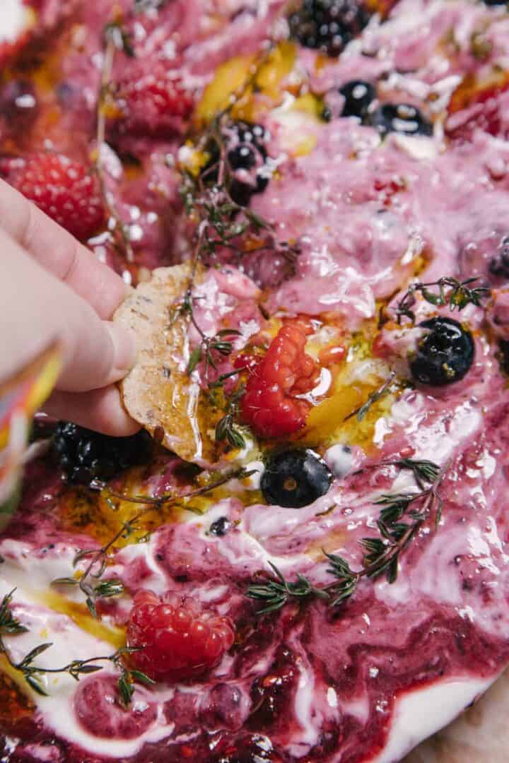 Close-up of a hand dipping a cracker into a creamy mixture topped with raspberries, blueberries, blackberries, olive oil, and sprigs of fresh thyme. The blend has a swirled appearance combining white and various berry colors.