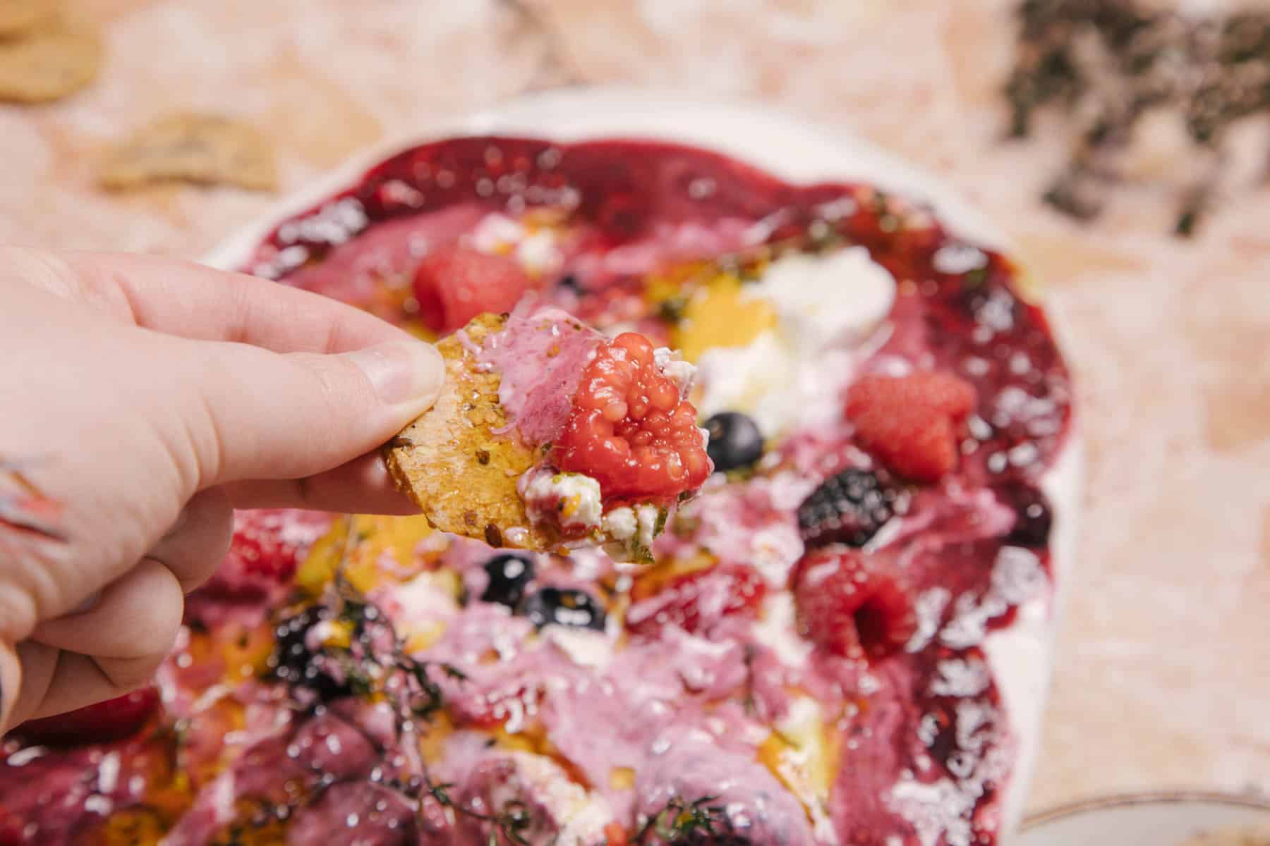 A hand holds a cracker topped with raspberry and a creamy pink spread. In the background, there's a colorful dish filled with various berries, including raspberries and blueberries, all set on a marbled surface.