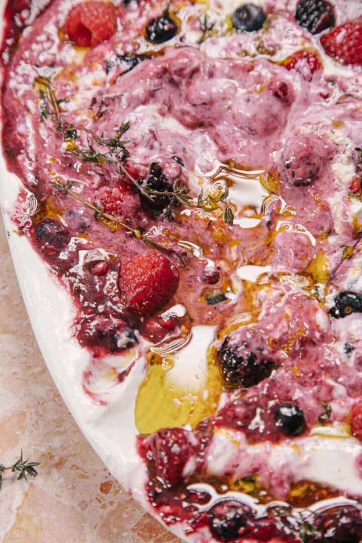 A close-up of a dish featuring a swirl of yogurt mixed with various berries, including raspberries, blackberries, and blueberries. The dish is drizzled with olive oil and garnished with fresh thyme, served on a white plate.