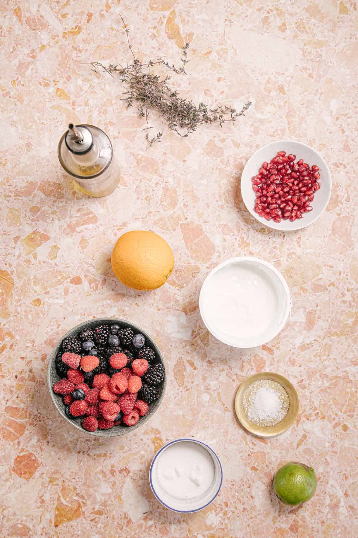 A top view showcases ingredients on a marble countertop: infused olive oil, fresh herbs, vibrant pomegranate seeds, mixed berries for berry labneh, a lemon, bowls of yogurt and sugar, salt in a small bowl, and a lime.