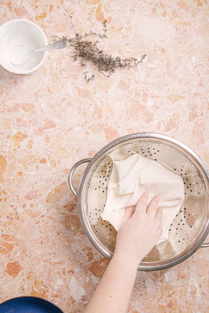 A hand places a white cloth inside a stainless steel colander on a marble countertop, preparing to craft berry labneh. Nearby, a small bowl with infused olive oil, a spoon, and some dried herbs add an aromatic touch to the scene.