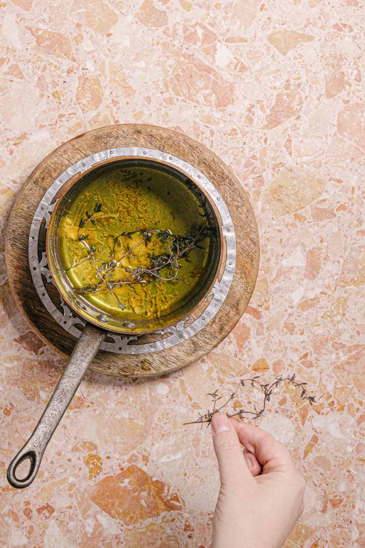 A hand holds herbs above a metallic pot of yellow liquid and herbs, on a wooden board. The countertop features a pink and beige speckled pattern, evoking the delicate hues of berry labneh drizzled with infused olive oil.