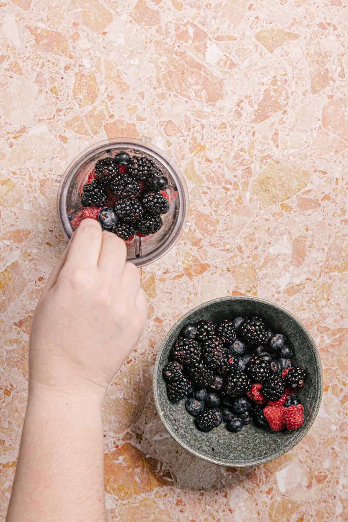 A hand placing mixed berries, including blackberries, blueberries, and raspberries, into a blender cup on a pink speckled countertop. A green bowl filled with the same types of berries is nearby.