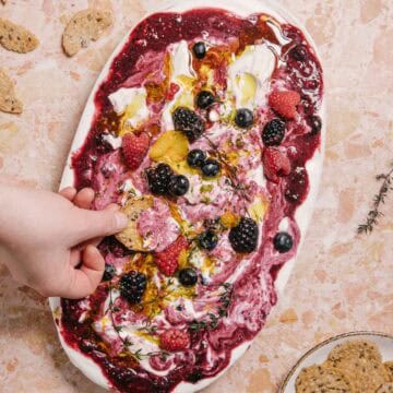 A hand dipping a cracker into a marble platter of yogurt swirled with berry sauce. Topped with raspberries, blackberries, blueberries, lemon zest, and thyme sprigs. Additional crackers and thyme are on the marble surface nearby.