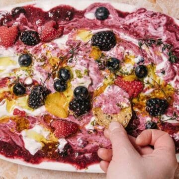 A hand dips a piece of bread into a colorful yogurt dip on an oval platter. The dip features swirls of deep purple and white, topped with raspberries, blueberries, blackberries, and herbs, set on a marbled surface.