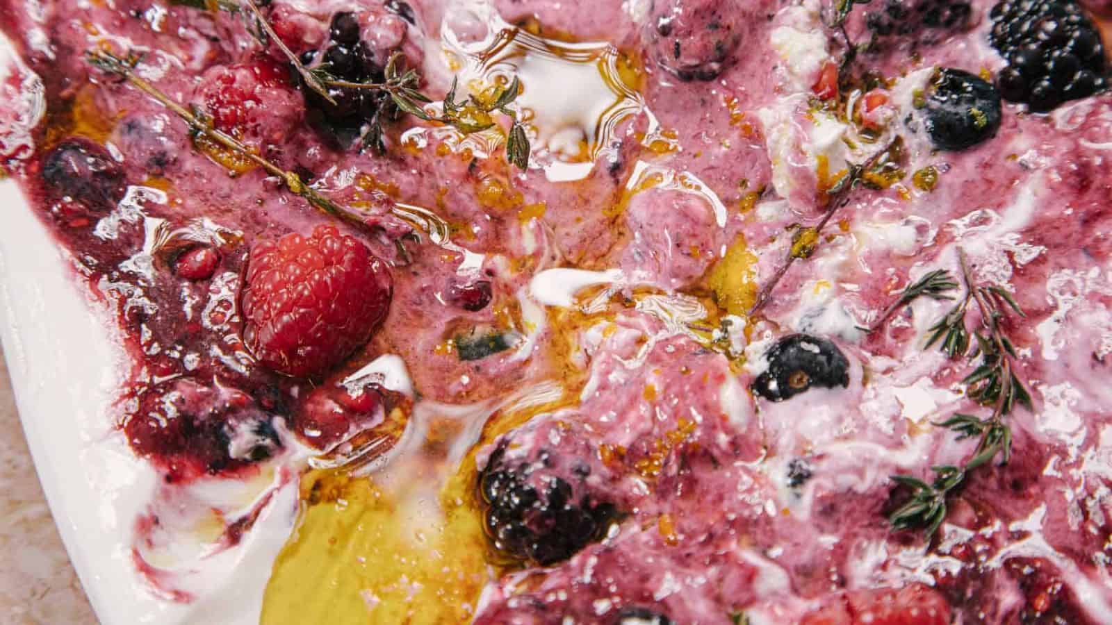 A hand dips a piece of bread into a colorful yogurt dip on an oval platter. The dip features swirls of deep purple and white, topped with raspberries, blueberries, blackberries, and herbs, set on a marbled surface.