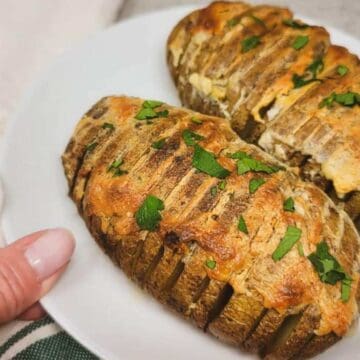 A person holds a white plate with two hasselback potatoes. The potatoes are sliced thinly and baked with a golden cheese topping, garnished with chopped herbs. A green-striped cloth is partially visible underneath.