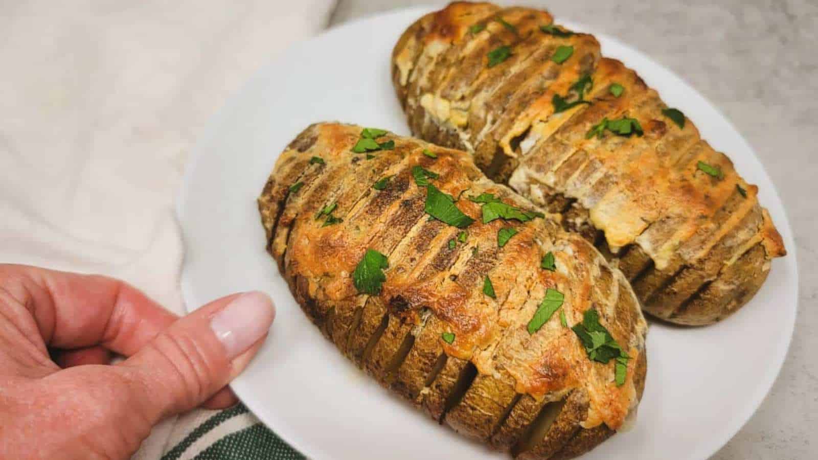 A person holds a white plate with two hasselback potatoes. The potatoes are sliced thinly and baked with a golden cheese topping, garnished with chopped herbs. A green-striped cloth is partially visible underneath.