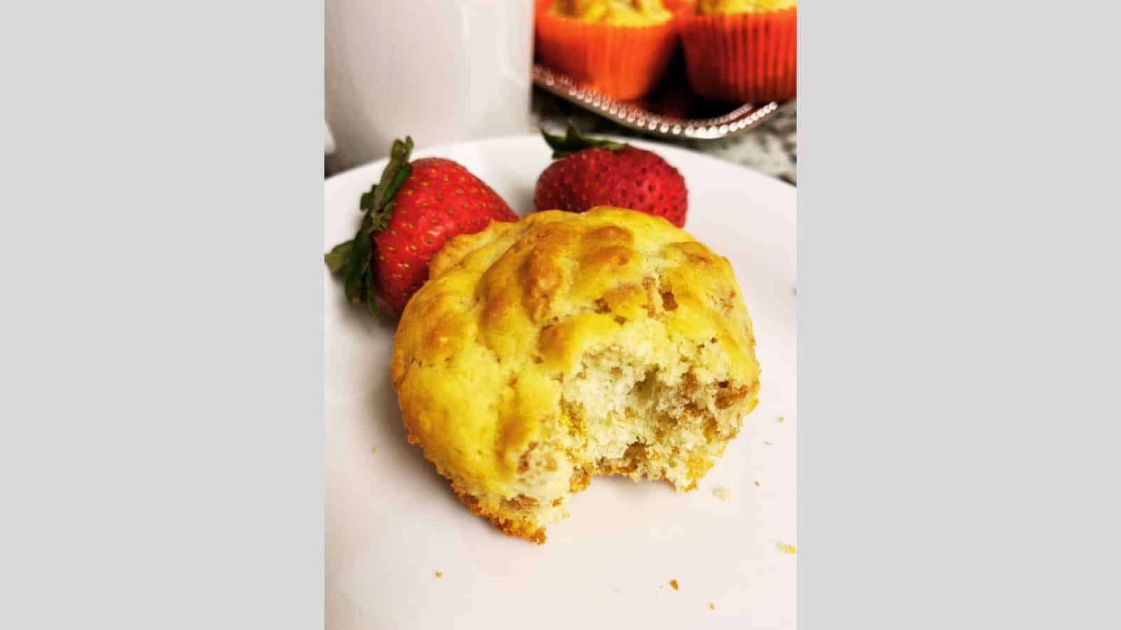 A bitten muffin rests on a white plate with two whole strawberries, embodying the essence of Grandma-Approved Dishes. In the background, a white mug and a tray with muffins in orange liners offer a taste like love from cherished recipes.