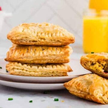 A stack of flaky pastries filled with scrambled ingredients is on a white plate. Two pastries reveal their filling. In the background, there are two glasses of orange juice and fresh berries in a white bowl. A striped cloth is nearby.