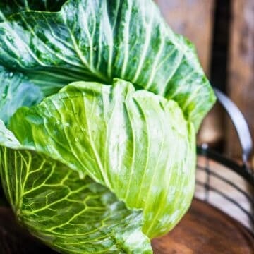 A fresh green cabbage with crisp leaves is placed on a wooden surface. The cabbage is whole and resting inside a round wire basket. The background features a rustic wooden texture with soft lighting.
