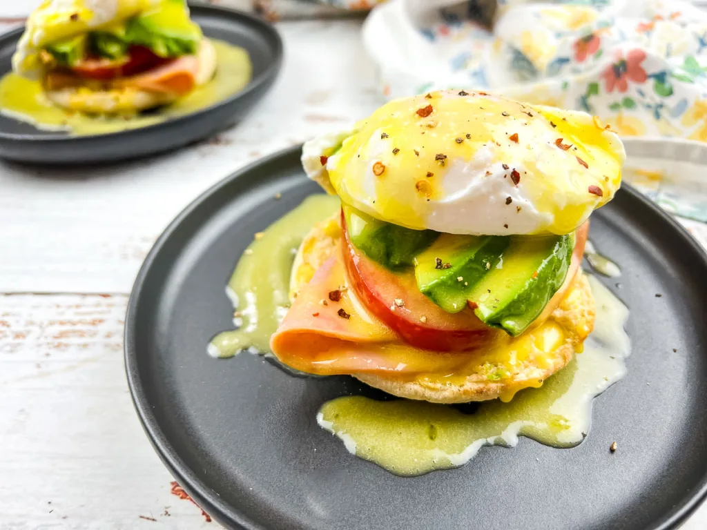 A plate of eggs benedict features a poached egg, avocado slices, ham, and a tomato slice on an English muffin, topped with hollandaise sauce and pepper flakes. Another similar dish is visible in the background.