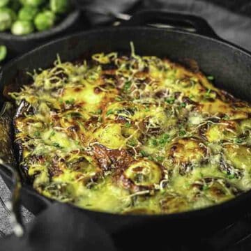 A cast iron skillet brimming with comforting casserole goodness, topped with melted cheese and herbs. The golden-brown surface reveals layers of vegetables, perfect for Sunday suppers. In the background, a small bowl of brussels sprouts completes this cozy comfort food scene.