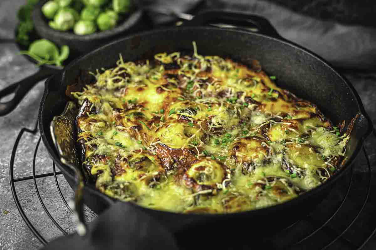 A cast iron skillet brimming with comforting casserole goodness, topped with melted cheese and herbs. The golden-brown surface reveals layers of vegetables, perfect for Sunday suppers. In the background, a small bowl of brussels sprouts completes this cozy comfort food scene.