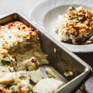 A baking dish filled with a baked dish, possibly a casserole, sits on a table. A portion has been removed and placed on a white plate beside it. The dish is topped with browned, melted cheese and specks of green herbs.