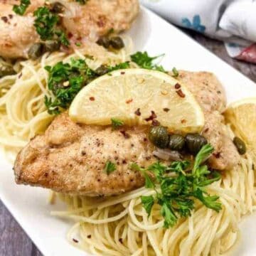 Two pieces of seasoned chicken are served on a bed of spaghetti, topped with capers, parsley, and red pepper flakes. Lemon slices garnish this classic dish presented on a white rectangular plate. A floral napkin is partially visible in the background, reminiscent of Grandma's table settings every time.