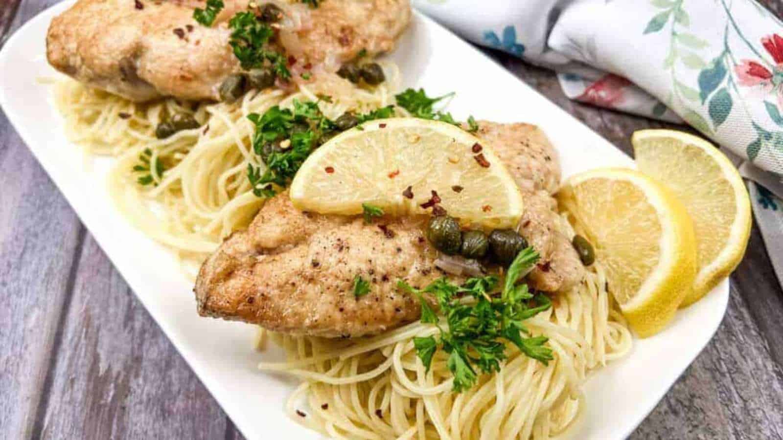 Two pieces of seasoned chicken are served on a bed of spaghetti, topped with capers, parsley, and red pepper flakes. Lemon slices garnish this classic dish presented on a white rectangular plate. A floral napkin is partially visible in the background, reminiscent of Grandma's table settings every time.