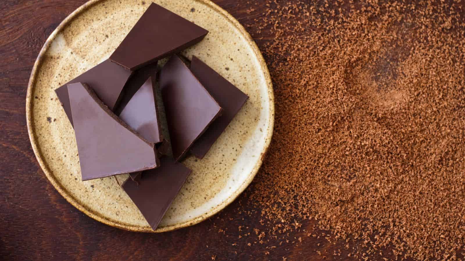 A beige plate with broken pieces of dark chocolate is on a wooden surface. Next to the plate, there's a scattering of grated chocolate powder on the table.