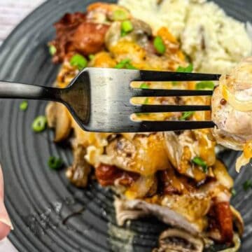 A fork holds a bite of chicken topped with onions and sauce. The plate in the background contains chicken with cheese and vegetables, rice, and garnished with chopped green onions. The scene suggests a ready-to-eat meal.
