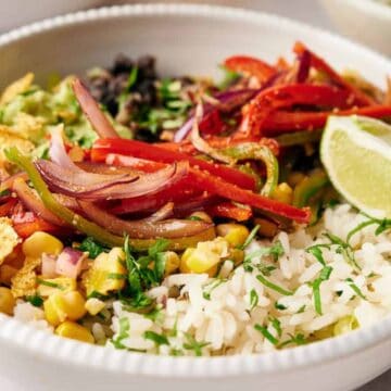 A bowl of rice topped with sliced red bell peppers, red onions, corn, black beans, chopped avocado, and tortilla strips makes for a family-friendly meal. With a lime wedge on the side and fresh herbs as garnish, it's perfect for tasty weeknight dinners.
