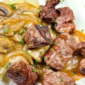 A plate of mashed potatoes topped with sliced mushrooms and brown gravy, accompanied by chunks of grilled beef. Chopped chives are sprinkled over the dish, making it a delightful nod to forgotten recipes of yesteryear.