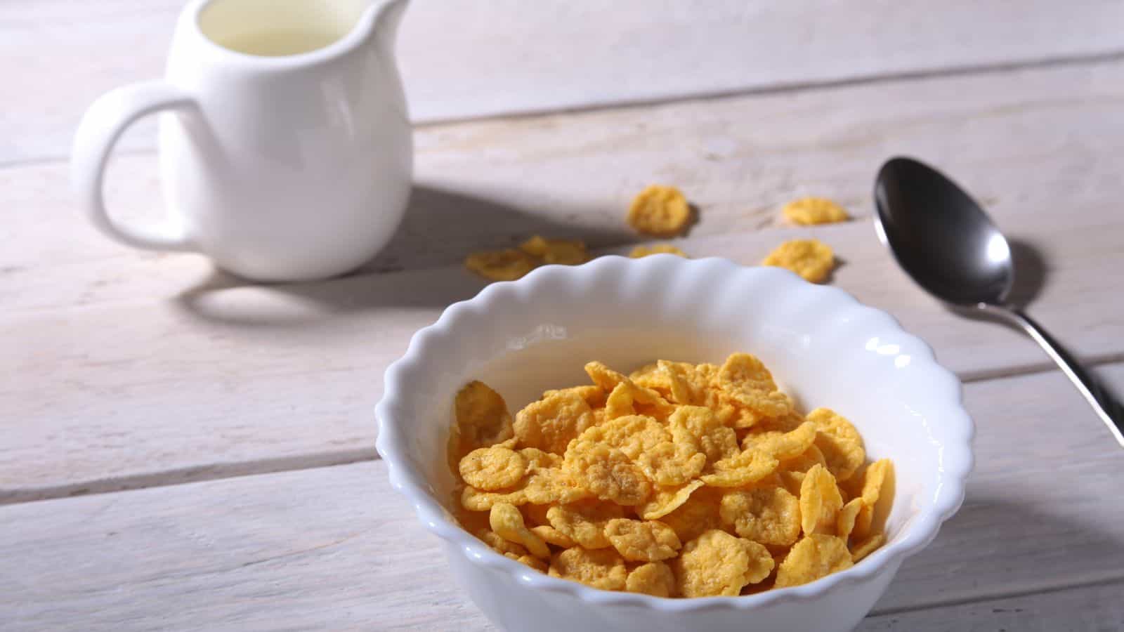 A white bowl filled with cornflakes sits on a wooden table. A few flakes are scattered around the bowl. Next to it, there is a metal spoon and a small white pitcher containing milk.