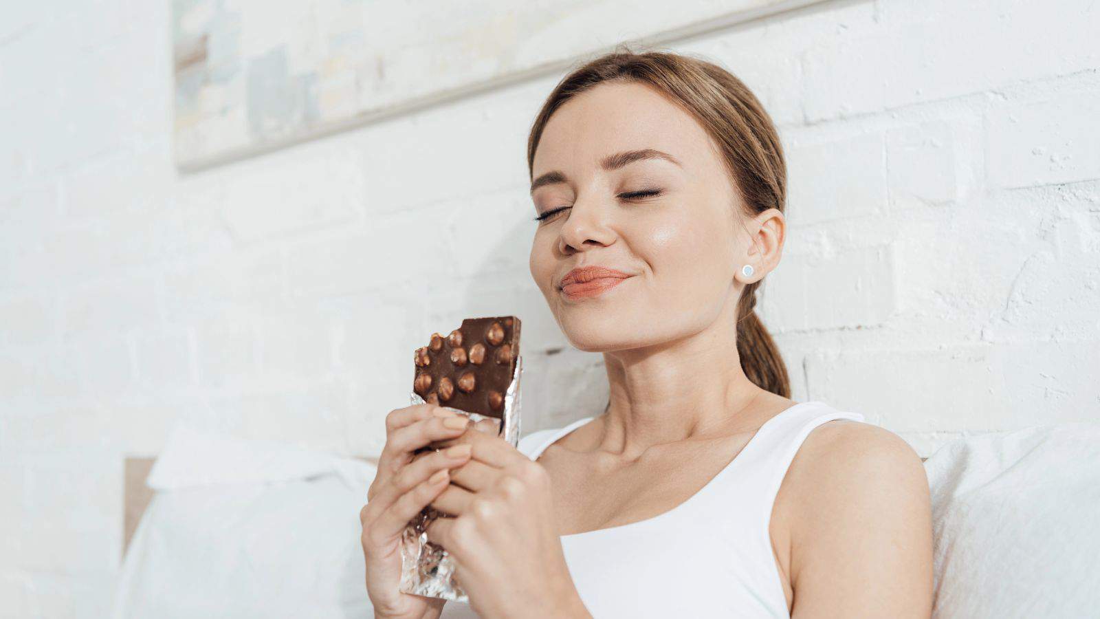 A woman in a white tank top is sitting against a white brick wall, holding a chocolate bar with nuts. Her eyes are closed, and she has a slight smile, appearing to savor the taste. A light-colored painting is in the background.