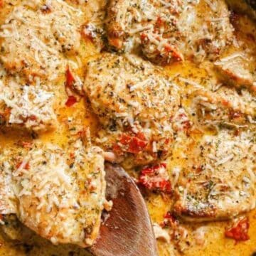 A skillet filled with chicken breasts in a creamy sauce, topped with grated cheese and herbs. Chunks of sun-dried tomatoes are visible in the sauce. A wooden spoon rests in the pan.