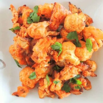 A plate of crispy buffalo cauliflower garnished with fresh parsley. The cauliflower pieces are coated in a spicy reddish sauce. A glass of red dipping sauce is visible in the background. Tongs and a sprig of parsley are beside the plate.