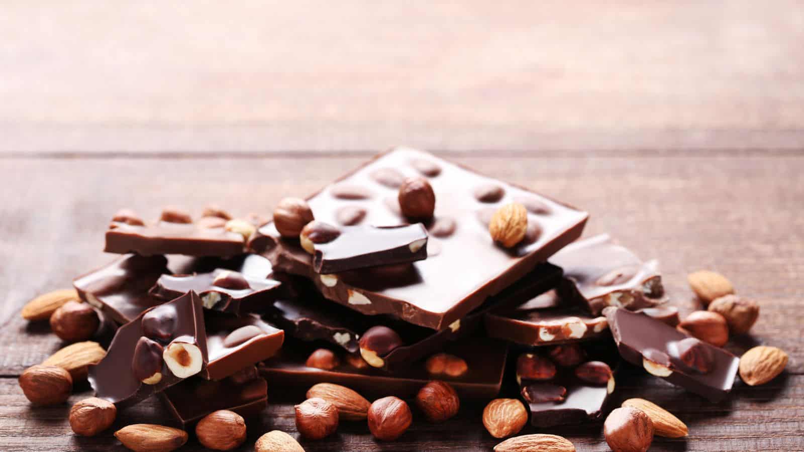 A stack of chocolate pieces is surrounded by scattered hazelnuts and almonds on a wooden surface. The chocolate pieces are embedded with whole nuts, creating a rustic and appetizing arrangement.