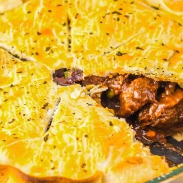 A pie with a golden-brown crust has a slice removed, revealing chunks of beef, peas, and possibly other vegetables in a thick sauce. The crust is neatly crimped around the edges and lightly sprinkled with herbs.