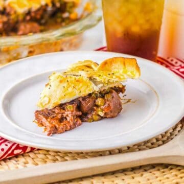 A slice of savory pie with a flaky crust and a visible filling of meat and vegetables sits on a white plate. The plate is on a red patterned cloth, with a wooden spoon nearby. A glass of iced tea and a full pie are in the background.