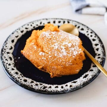 A serving of sweet potato casserole topped with powdered sugar sits on a black ceramic plate with a speckled pattern. Beside it, a gold spoon rests elegantly. In the background, a folded cloth napkin peeks through, reminiscent of savoring gluten-free carrot soufflé delights.