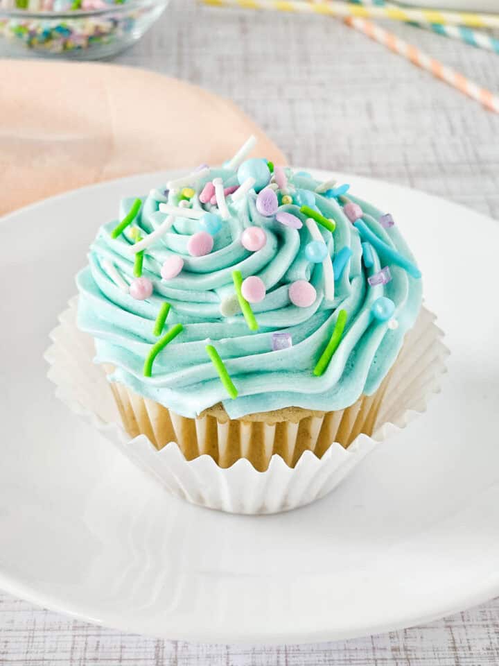 A gluten-free vanilla cupcake with light blue frosting sits on a white plate. The frosting is topped with colorful sprinkles in green, pink, and purple. The cupcake is wrapped in a white paper liner, while a blurred background features a napkin and striped straws.