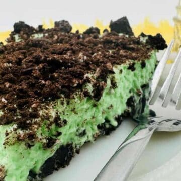 A slice of dessert on a white plate features a bright green creamy layer sprinkled with crumbled chocolate cookies, reminiscent of childhood sweet treats. A fork rests on the plate, inviting a taste. A jar containing more crumbled cookies is partially visible in the background.