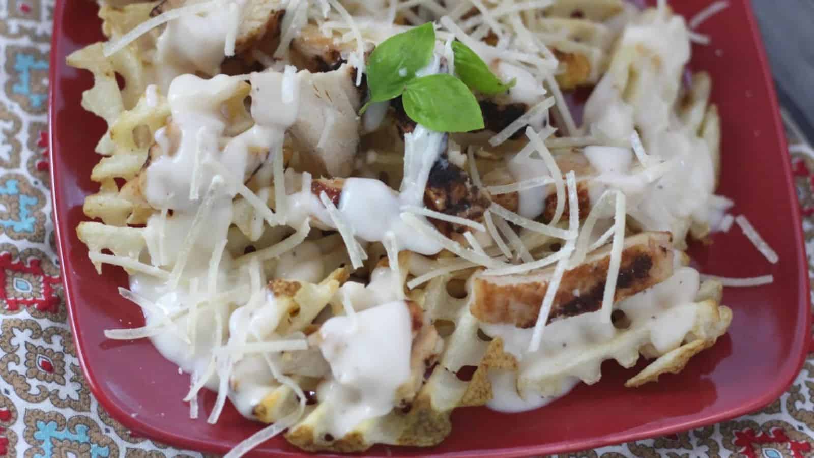 A red plate brimming with waffle fries topped with grilled chicken pieces, shredded cheese, and drizzled with a creamy sauce—perfect for those who love grilled meals. A small green basil garnish rests on top, embodying the essence of a Backyard BBQ on a patterned surface.
