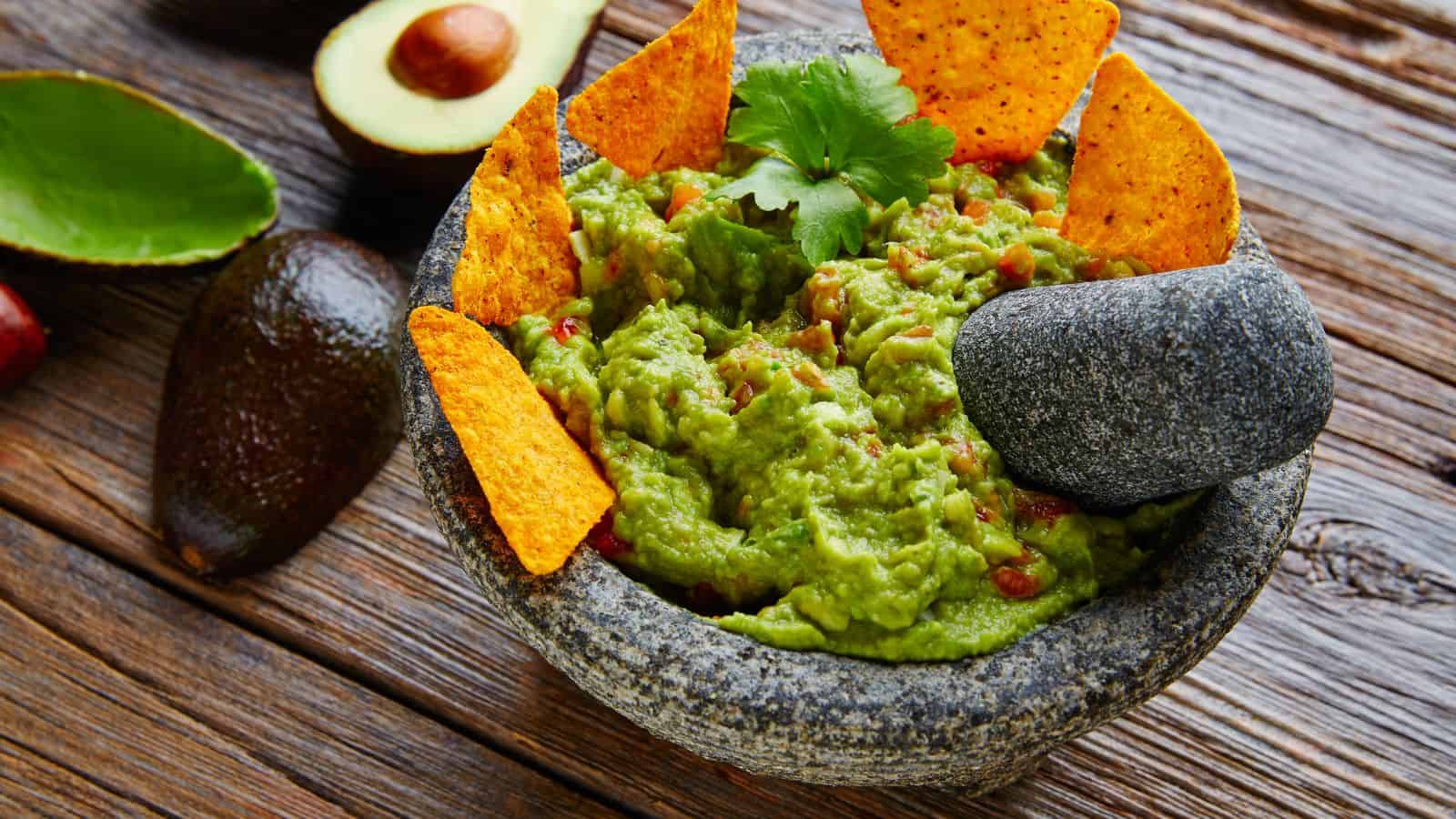 A stone mortar filled with guacamole, garnished with tortilla chips and a cilantro leaf. A stone pestle is resting on the side. Avocado halves and a seed are visible on the wooden table beside it.