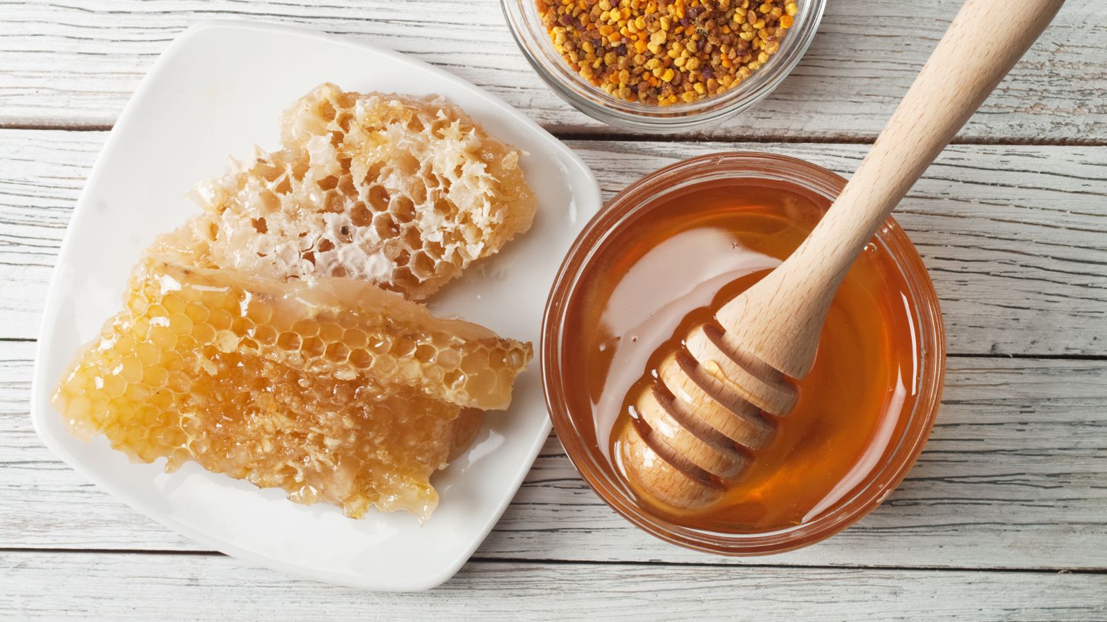 A white plate with honeycomb sits next to a glass jar of honey containing a wooden honey dipper. A small bowl of bee pollen granules is nearby on a white wooden surface.