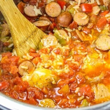 A wooden spoon stirs a vibrant one-pot meal in a pot, brimming with sliced sausages, diced tomatoes, chopped green peppers, onions, and grains of rice. The simple dish simmers away with visible steam rising from the flavorful liquid.