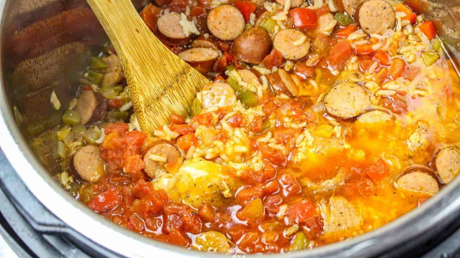 A wooden spoon stirs a vibrant one-pot meal in a pot, brimming with sliced sausages, diced tomatoes, chopped green peppers, onions, and grains of rice. The simple dish simmers away with visible steam rising from the flavorful liquid.