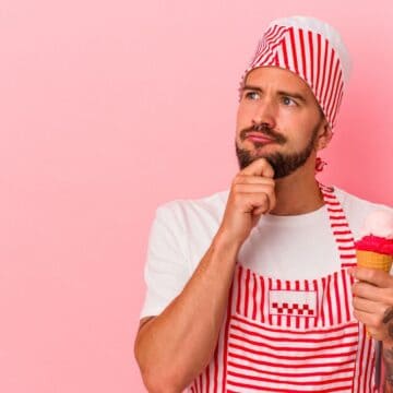 A man wearing a red and white striped apron and hat holds an ice cream cone with pink scoops. He has a thoughtful expression, with one hand on his chin. The background is a solid pink color.
