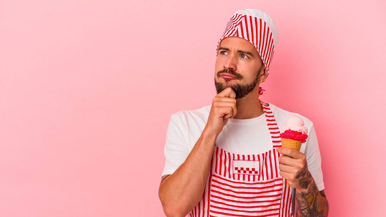 A man wearing a red and white striped apron and hat holds an ice cream cone with pink scoops. He has a thoughtful expression, with one hand on his chin. The background is a solid pink color.