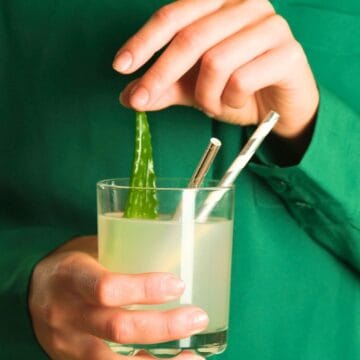 A person in a green shirt holds a glass of green liquid with a clear wedge, possibly aloe vera. Two striped straws are in the glass. The person is using their hand to adjust the wedge.