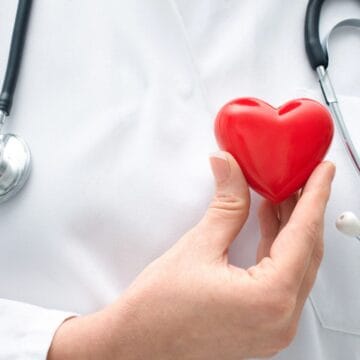 A person wearing a white coat and a stethoscope holds a red heart-shaped object in their hand.