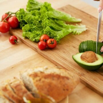 A person slices an avocado on a wooden cutting board. Nearby are cherry tomatoes and leafy lettuce. In the foreground, there is a loaf of bread. The setting is a clean, light-colored kitchen surface.