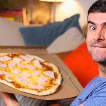 A man in a blue shirt is sitting on a couch, holding an open pizza box with a pizza topped with slices of ham and pineapple. He is looking at the camera with a surprised expression. There are cushions and a plant in the background.