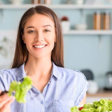 A woman with long brown hair and a light blue shirt is smiling in a kitchen while holding a bowl of salad in one hand and a fork with lettuce in the other. Shelves and a refrigerator are visible in the background.